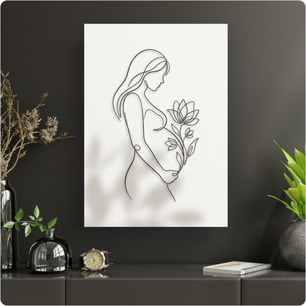 Affiche murale minimaliste représentant une femme enceinte de profil, les mains posées sur son ventre arrondi, tenant délicatement une fleur de lotus en pleine éclosion. Le dessin est réalisé en une seule ligne noire continue sur un fond blanc texturé, créant une ombre douce. L'affiche est présentée dans un intérieur moderne aux murs gris foncé, posée sur un meuble bas avec des objets décoratifs comme un vase et une plante verte.
