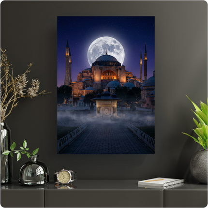 Affiche verticale représentant la majestueuse Sainte-Sophie à Istanbul sous une pleine lune éclatante et un ciel étoilé profond. Le monument historique est illuminé de lumières chaudes, avec ses minarets élancés. Un chemin pavé mène à une fontaine ancienne, entourée d'une légère brume au premier plan, créant une atmosphère mystique et sereine. L'affiche est présentée sur un mur gris foncé dans un intérieur moderne, avec des éléments décoratifs végétaux et un réveil.