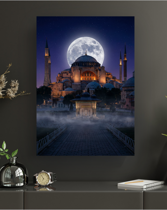 Affiche verticale représentant la majestueuse Sainte-Sophie à Istanbul sous une pleine lune éclatante et un ciel étoilé profond. Le monument historique est illuminé de lumières chaudes, avec ses minarets élancés. Un chemin pavé mène à une fontaine ancienne, entourée d'une légère brume au premier plan, créant une atmosphère mystique et sereine. L'affiche est présentée sur un mur gris foncé dans un intérieur moderne, avec des éléments décoratifs végétaux et un réveil.