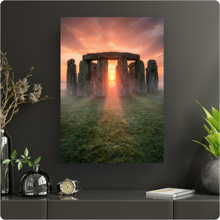 Affiche verticale de Stonehenge au lever du soleil. Les imposantes pierres grises du monument mégalithique sont baignées dans une lumière orange et rose intense émanant du soleil bas, perçant à travers les arches et la brume légère au sol. Le ciel est teinté de couleurs chaudes, contrastant avec l'herbe verte et humide au premier plan. L'ensemble dégage une atmosphère mystique et majestueuse.