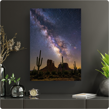 Affiche verticale capturant la majesté de la Voie Lactée au-dessus des formations rocheuses emblématiques de Monument Valley. Le ciel nocturne est un spectacle d'étoiles scintillantes et de nébuleuses aux teintes violettes, roses et bleues. Au premier plan, plusieurs grands cactus saguaro se dressent fièrement dans le paysage désertique, éclairé par une douce lueur orangée à l'horizon. Une étoile filante traverse le ciel, ajoutant une touche de magie à cette scène grandiose.